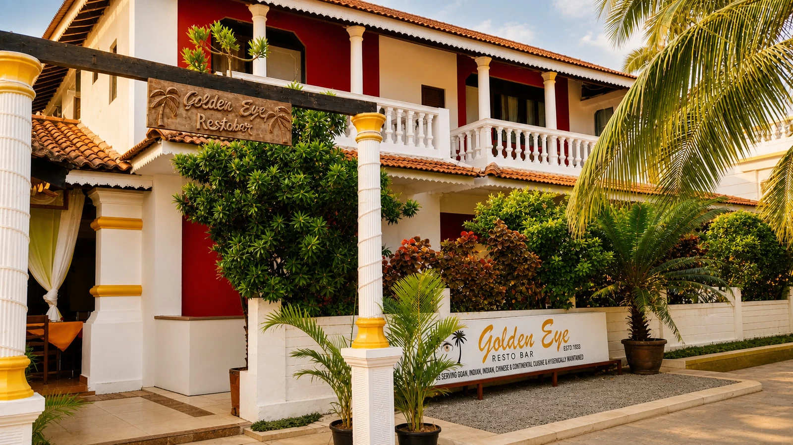 Hotel Golden Eye at golden hour: the red and white house behind its garden, white columns, terracotta roof and coconut palms.